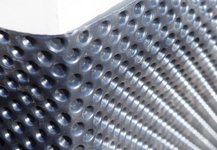 Nahaufnahme einer strukturierten Metall- oder Kunststoffoberfläche mit gleichmäßig verteilten, erhabenen runden Noppen, die entlang einer Kante ein Gittermuster bilden. Die Beleuchtung unterstreicht die glänzende, reflektierende Beschaffenheit des Materials.