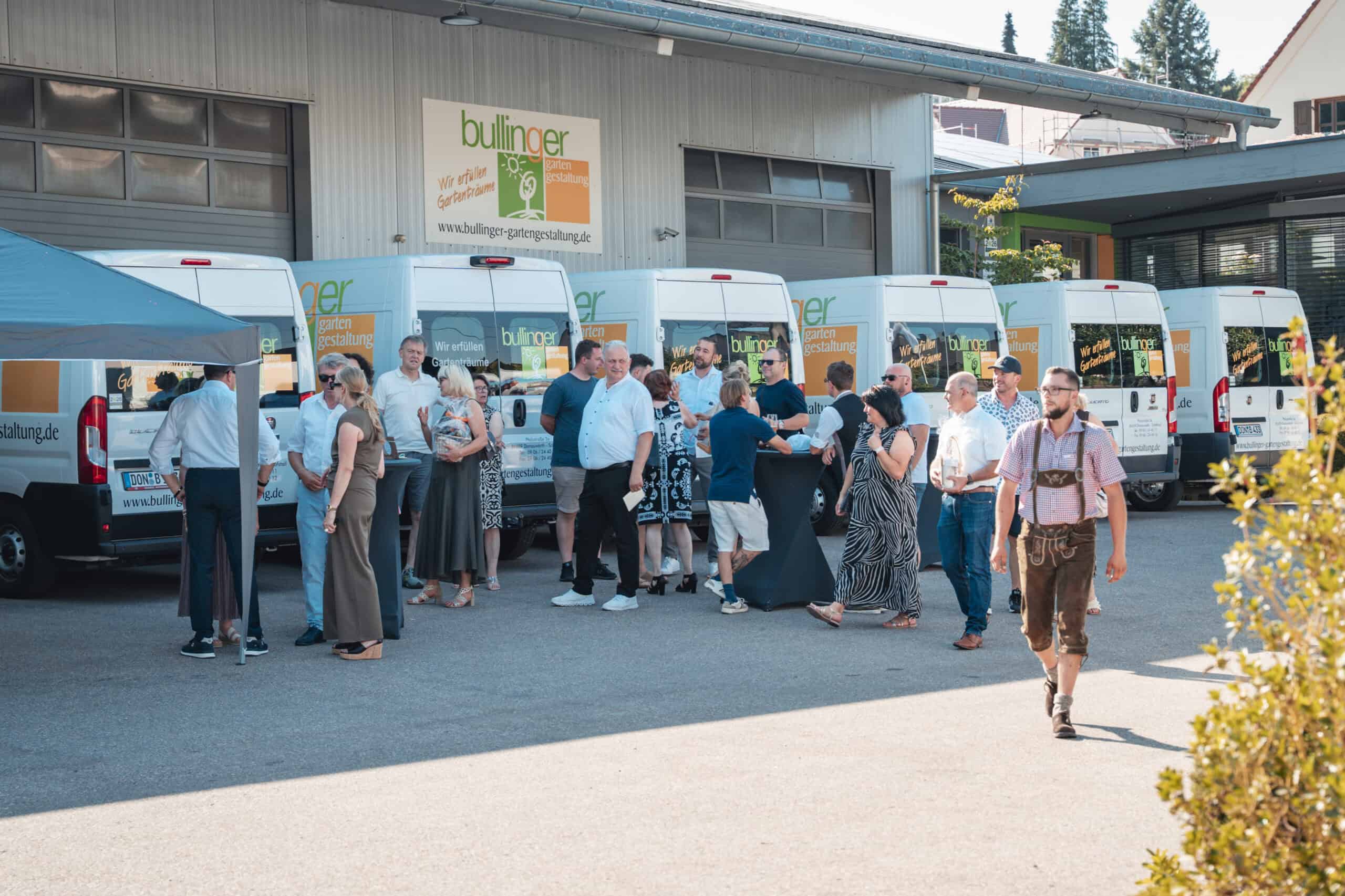 Eine Gruppe von Menschen versammelt sich und unterhält sich in der Nähe mehrerer weißer Transporter, die vor einem Gebäude geparkt sind, während im Vordergrund ein Mann in traditioneller Kleidung vorbeigeht.